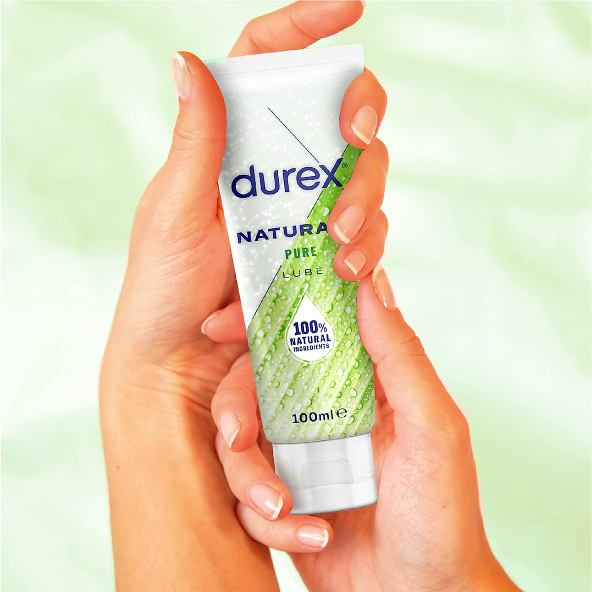 A pair of hands holding the 100ml tube of Durex Naturals Pure Lube against a blurred, light green background, providing a sense of the product's scale and texture.