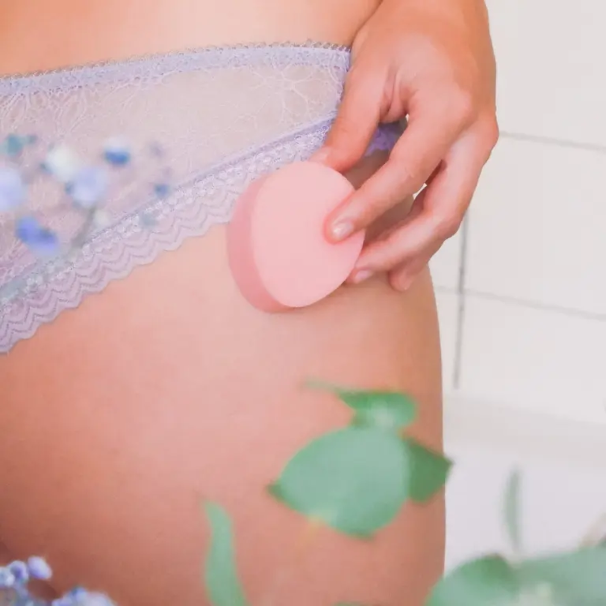 A lifestyle photograph showing a person's hand holding a pink sponge tampon against their upper thigh. The person is wearing light purple lace underwear. The background is soft-focused with hints of green leaves and white tiles, suggesting a bathroom or personal care setting.