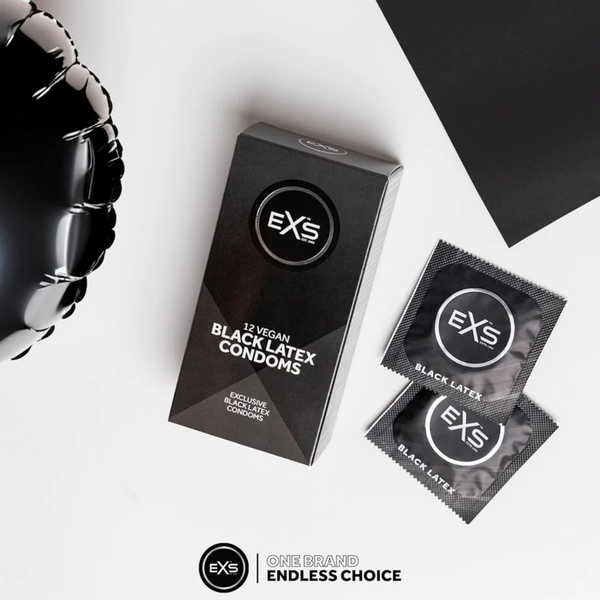 Overhead flat lay photo of the EXS 12 Vegan Black Latex Condoms package sitting next to two individual foil wrappers on a clean white surface. A shiny black balloon is visible in the corner.