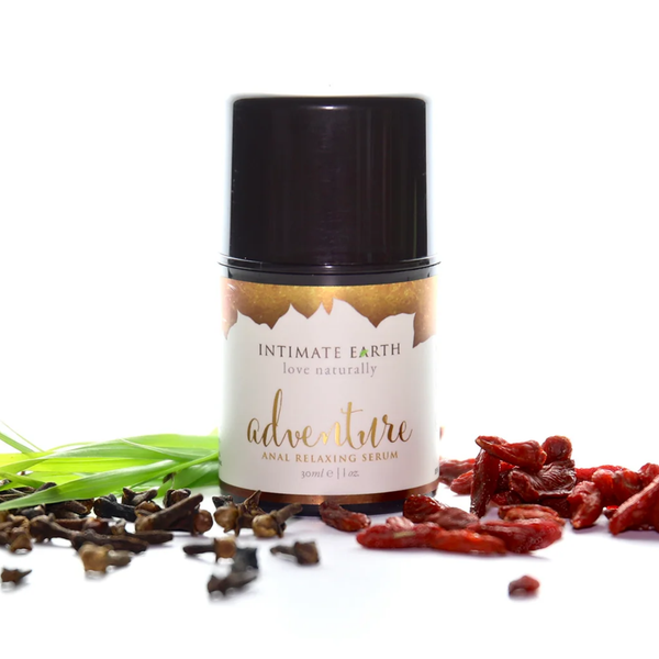 Nature-inspired studio photo showing the 30 ml (1 fl oz) bottle of Intimate Earth Adventure anal relaxing serum surrounded by fresh green grass, whole cloves, and dried goji berries.