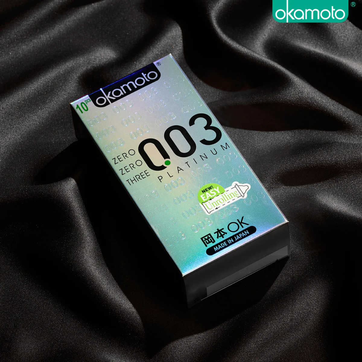 A shot of a Okamoto 003 Platinum 10-piece box resting on luxurious black silk fabric. The box features a holographic silver finish with the signature '003' logo. A bright green sticker on the front highlights the "New! Easy Unrolling" feature.
