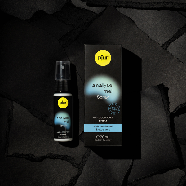 A product shot displays a black and white bottle of pjur "analyse me! Spray" next to its matching black box, both resting on a textured dark gray surface. 