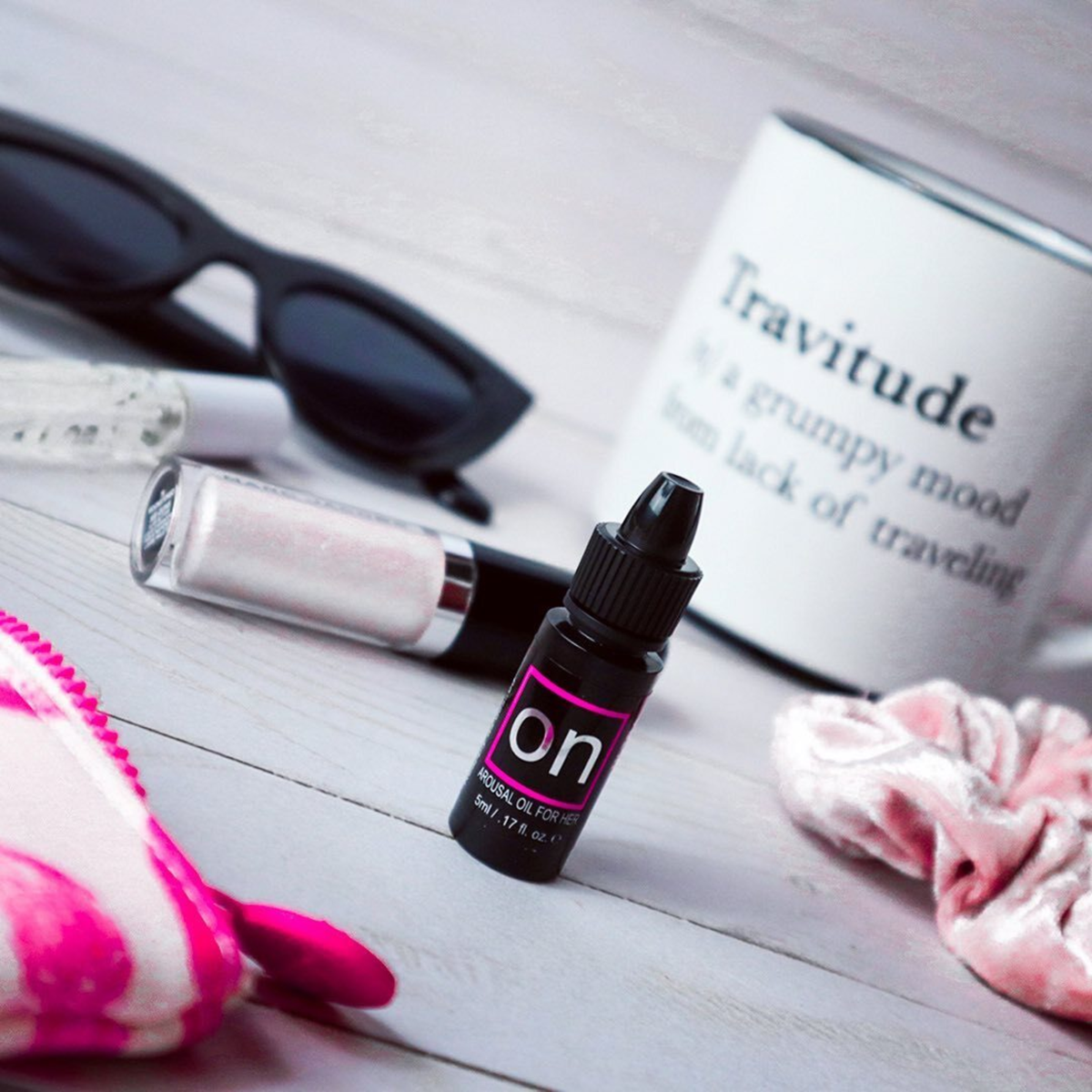 A flat-lay lifestyle shot featuring a miniature bottle of Sensuva ON Arousal Oil standing upright on a wooden desk. A white mug reading "Travitude," sunglasses, and a pink scrunchie sit softly blurred in the background.