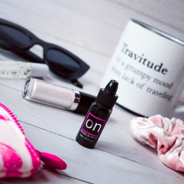 A flat-lay lifestyle shot featuring a miniature bottle of Sensuva ON Arousal Oil standing upright on a wooden desk. A white mug reading "Travitude," sunglasses, and a pink scrunchie sit softly blurred in the background.