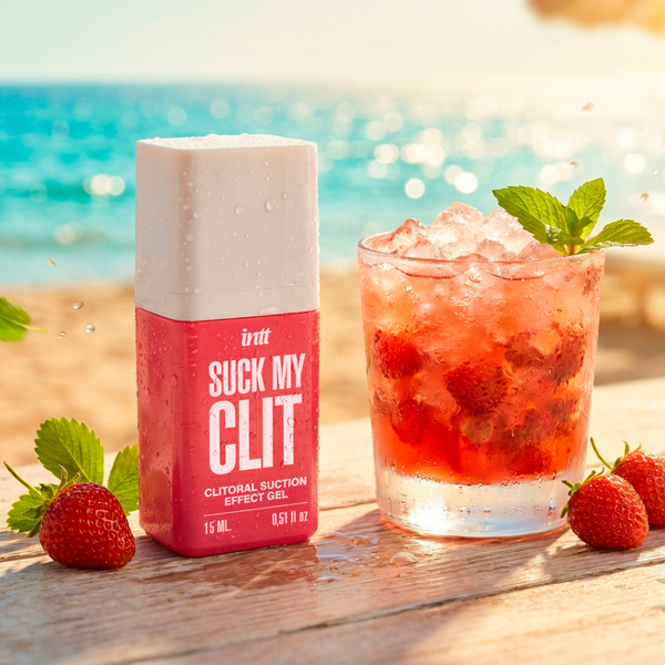 The pink intt Suck My Clit Wild Strawberry product bottle stands on a weathered wooden table next to a refreshing, iced strawberry cocktail garnished with mint. A sunny beach and blue ocean serve as the background.