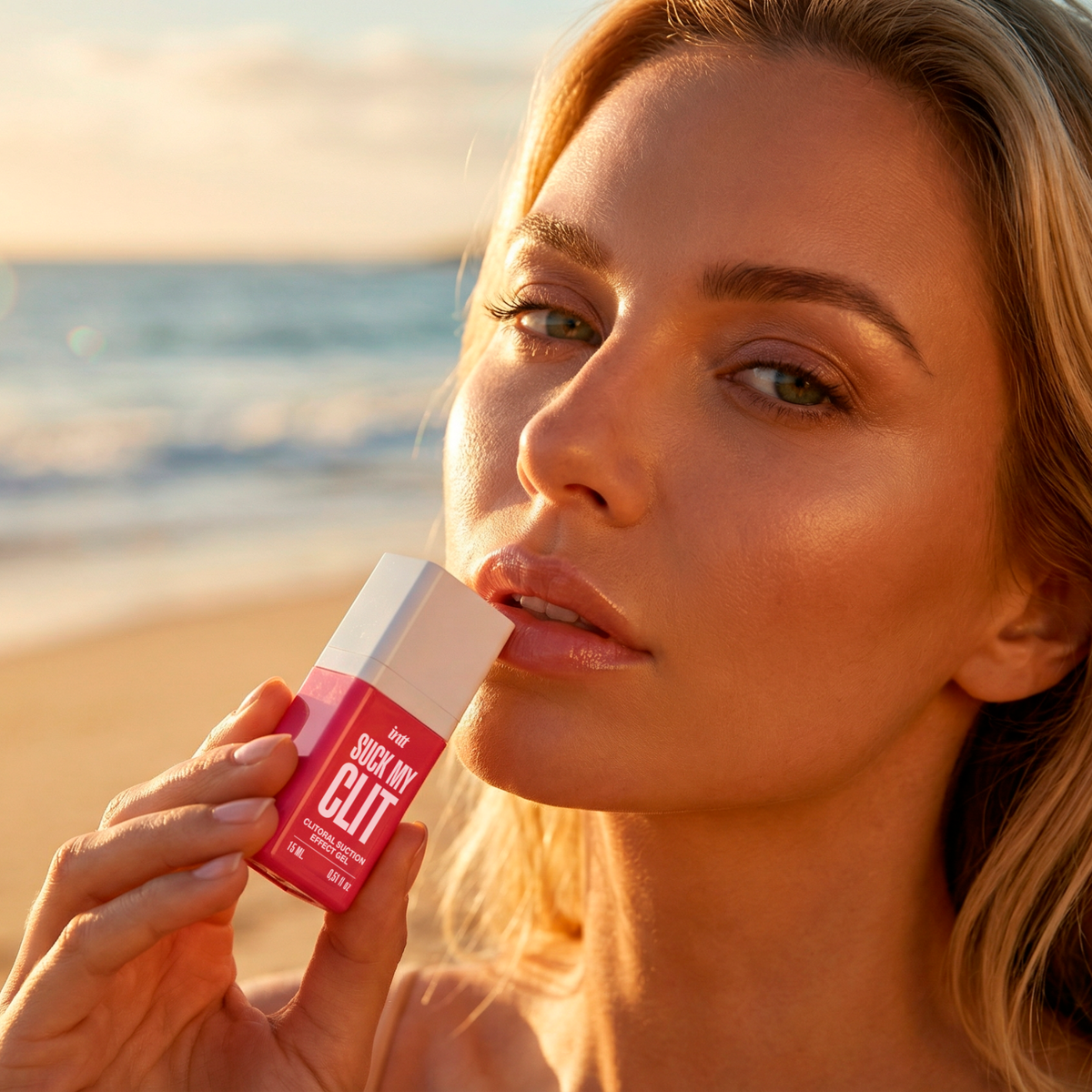 A close-up portrait of a person with blonde hair holding the pink intt Suck My Clit Wild Strawberry product bottle up to their lips, with a soft-focus beach and ocean sunset in the background.