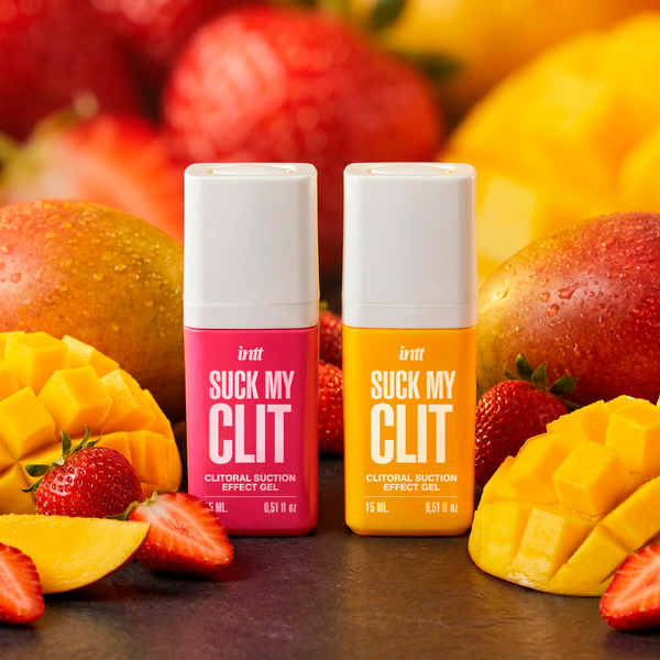 Two intt Suck My Clit product bottles stand side-by-side on a dark surface: the hot pink Wild Strawberry version and the bright yellow Tropical Mango version, beautifully surrounded by a mix of fresh strawberries and sliced mangoes.
