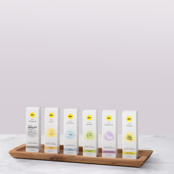 Six white boxes of pjur med personal lubricant in a wooden tray on a marble surface against a light purple background. The boxes are labeled "PREMIUM," "SOFT," "NATURAL," "REPAIR," "SENSITIVE," and "VEGAN."