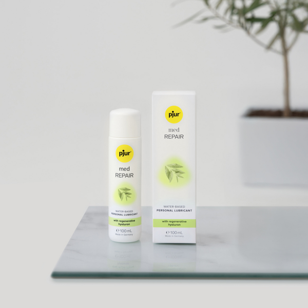 A white bottle and box of pjur med REPAIR water-based personal lubricant stand on a light grey marble surface with a blurred potted plant in the background.