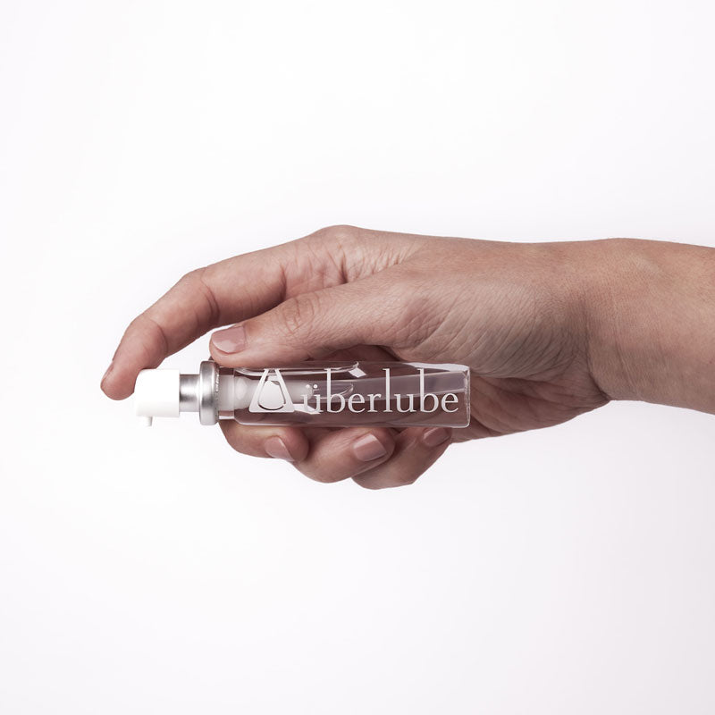 A hand holds a clear, horizontal Überlube lubricant spray bottle, prominently displaying the "überlube" logo. This compact WorldCondoms product offers discreet and convenient personal lubrication.