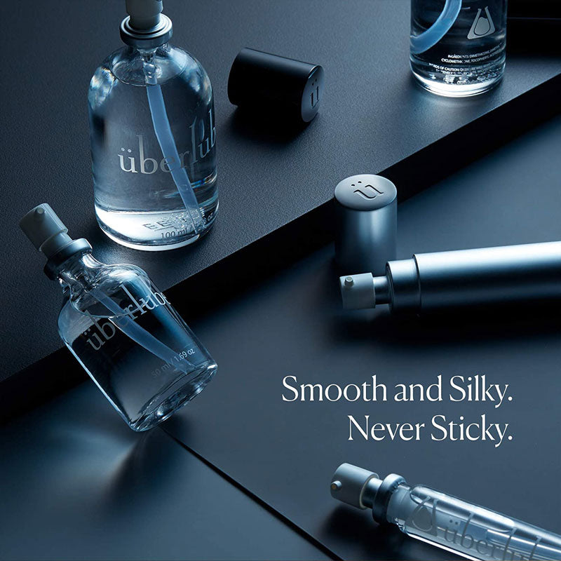 This close-up shot features multiple clear glass Überlube lubricant bottles, some with their caps off, arranged on a dark, sleek surface. The text "Smooth and Silky. Never Sticky." is prominent, highlighting a key benefit of this WorldCondoms lubricant.
