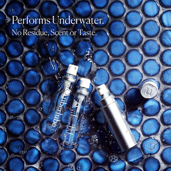 Three Überlube lubricant bottles, two clear glass and one silver, lie amidst dark blue, circular, tile-like bubbles filled with water. The text "Performs Underwater. No Residue, Scent or Taste." highlights key features of this WorldCondoms lubricant.
