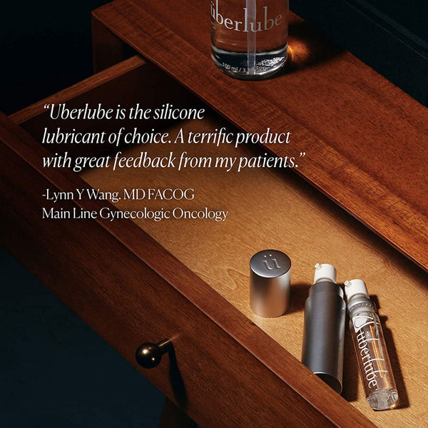 A partial view of a wooden dresser drawer features three Überlube products: a silver cap, a silver spray bottle, and a clear glass spray bottle with the "überlube" logo. A quote from Lynn Y. Wang, MD FACOG, praises Überlube as "the silicone lubricant of choice" with "terrific product with great feedback from my patients," underscoring WorldCondoms' commitment to quality and trusted products.
