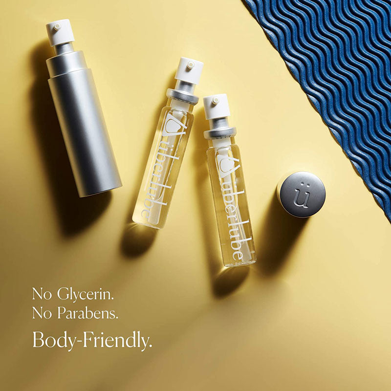 Three Überlube bottles—one silver and two clear glass—are artfully arranged on a two-toned background of yellow and textured blue. The text "No Glycerin. No Parabens. Body-Friendly." emphasizes the safe and thoughtful formulation of this WorldCondoms lubricant.